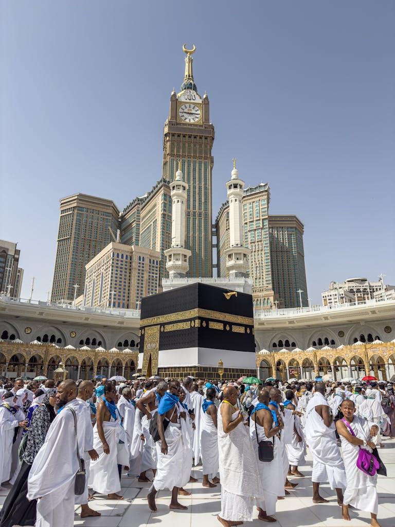 A large group of pilgrims gather at the Kaaba in Mecca, Saudi Arabia, during the Hajj pilgrimage under a clear blue sky.